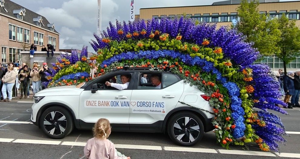 Een witte SUV, bedekt met regenboogbloemen, rijdt mee in een straatparade, met mensen en een kind in de buurt.