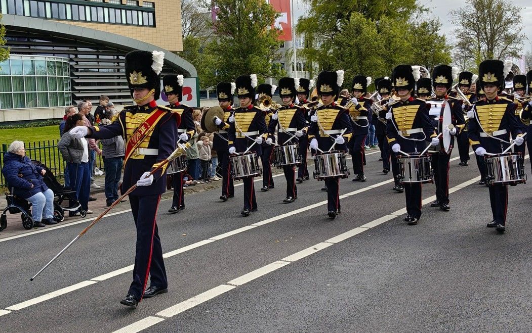Een fanfarekorps in marineblauwe uniformen met hoeden marcheert door een straat langs toeschouwers.