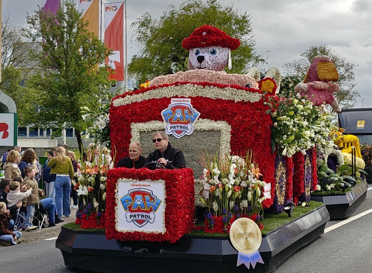 Een met bloemen bedekte praalwagen met een rode brandweerwagen als voorkant en een dalmatiër als mascotte.
