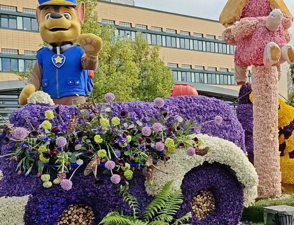 Bloemensculptuur van een gele cartoonhond in een blauw uniform op een paars, met bloemen versierd voertuig.