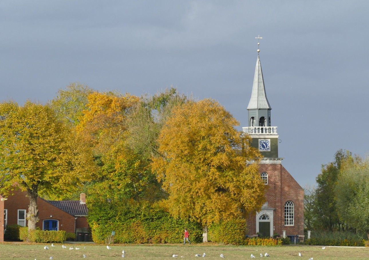 Kerk met hoge torenspits, omgeven door bomen met geel en oranje herfstblad.