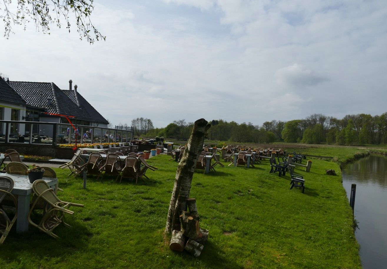Een woning aan het meer met een wit gebouw, een groen gazon, tafels en een afgezaagde berkenboomstam. Bewolkte lucht.