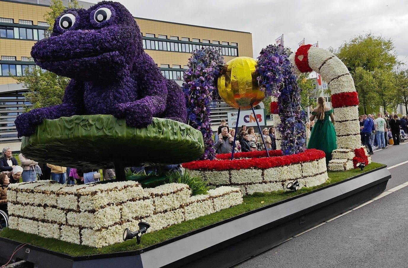 Paarse cartoonfiguur met dieren en kleurrijke bloemen op een praalwagen, met een rood-witte boog en mensen aan boord.