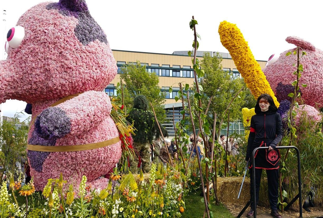 Roze bloemsculpturen in een tuin met een persoon ernaast en een gele bloemenboog erachter.