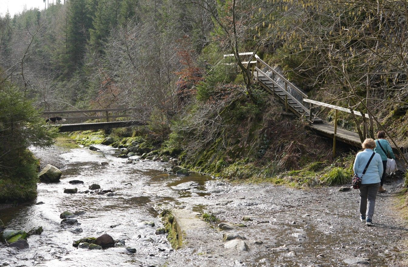 Twee mensen lopen over een rotsachtig pad langs een ondiepe beek, met een houten loopbrug en een trap zichtbaar op de oever.