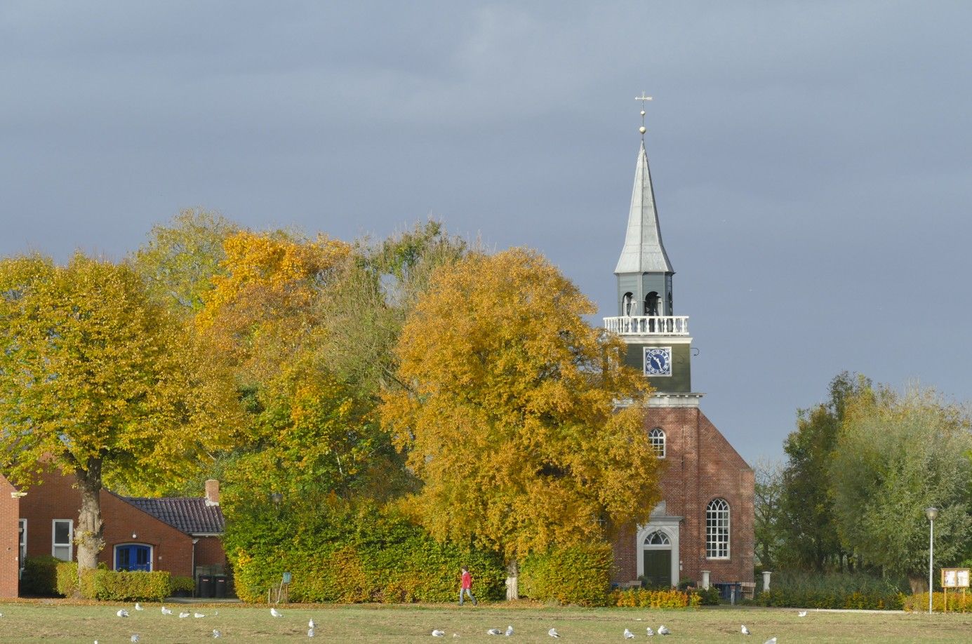 Een bakstenen kerk met een torenspits, omgeven door bomen met gele en groene bladeren, onder een bewolkte hemel.