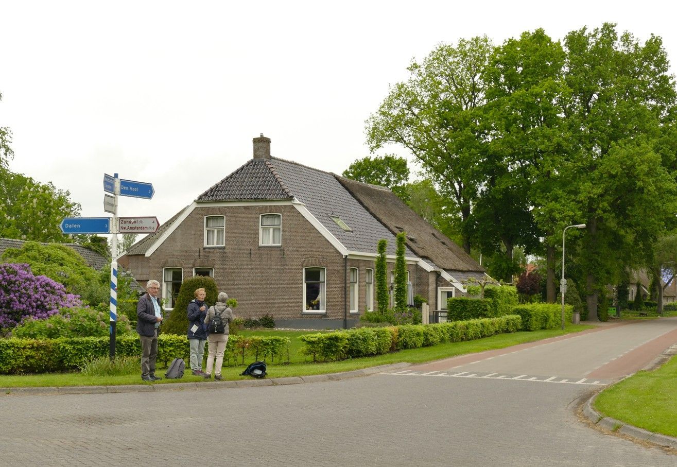 Een bakstenen huis met een donker dak en groene hagen; mensen staan ​​bij een wegwijzer langs de weg.