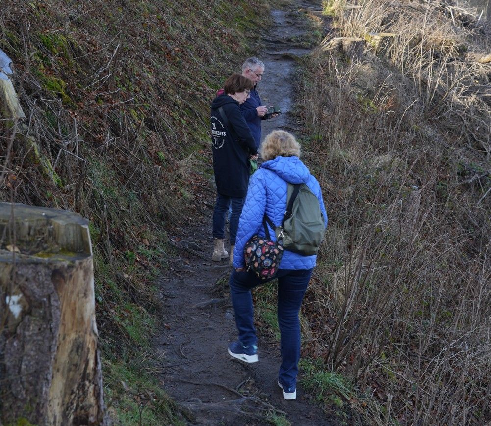 Drie mensen lopen over een smal, onverhard wandelpad door een bosrijk gebied met droge begroeiing en een boomstronk in de buurt.