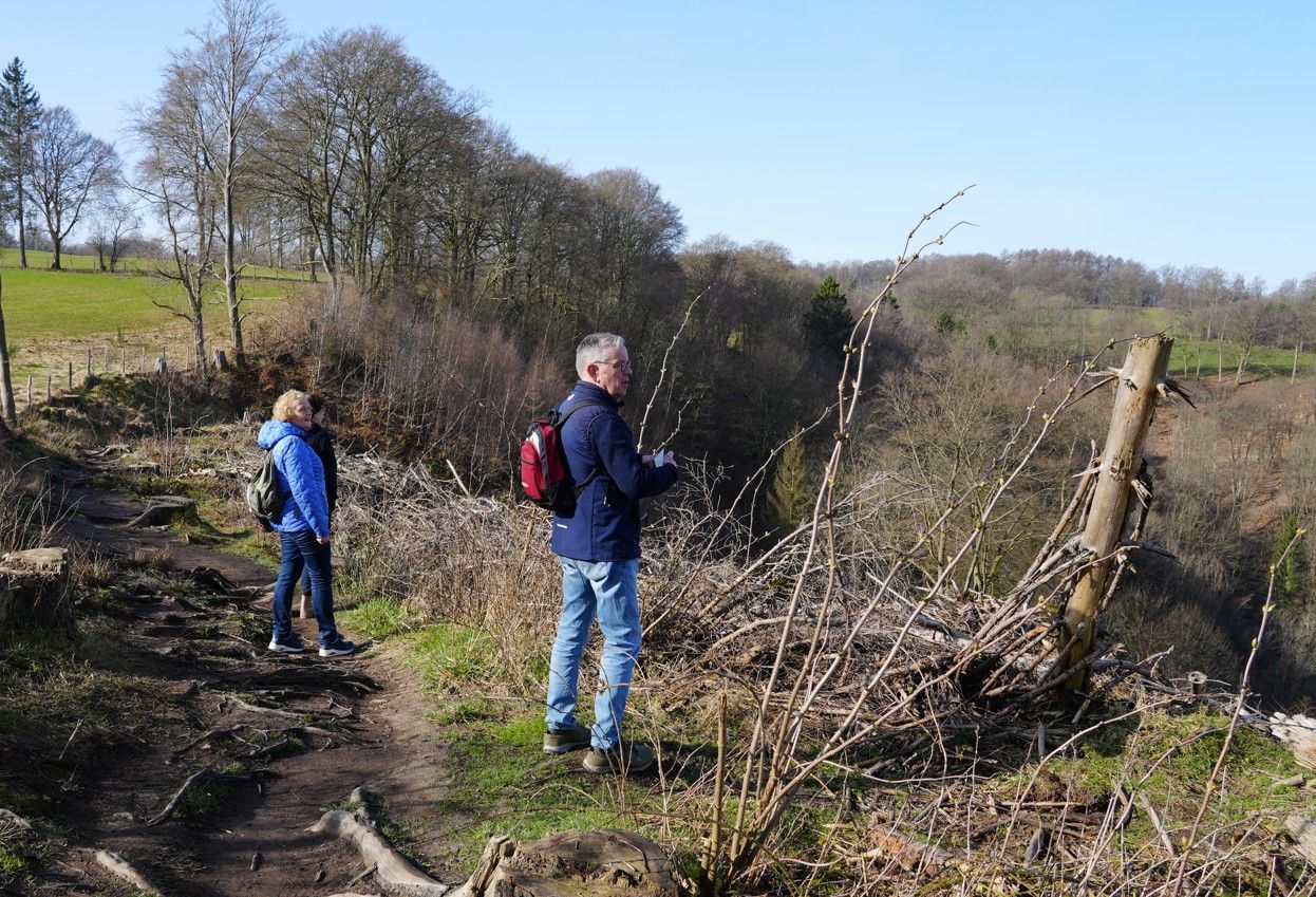 Twee mensen in casual buitenkleding staan op een zandpad met uitzicht op een beboste vallei op een heldere, zonnige dag.