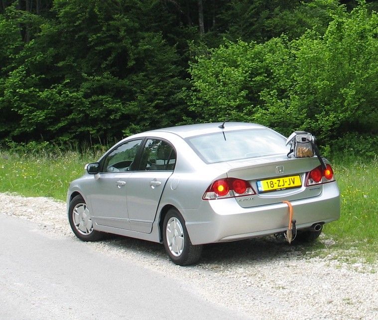 Een zilverkleurige auto staat geparkeerd langs de weg, met een rugzak op de achterklep. Op de achtergrond zijn groene bomen te zien.