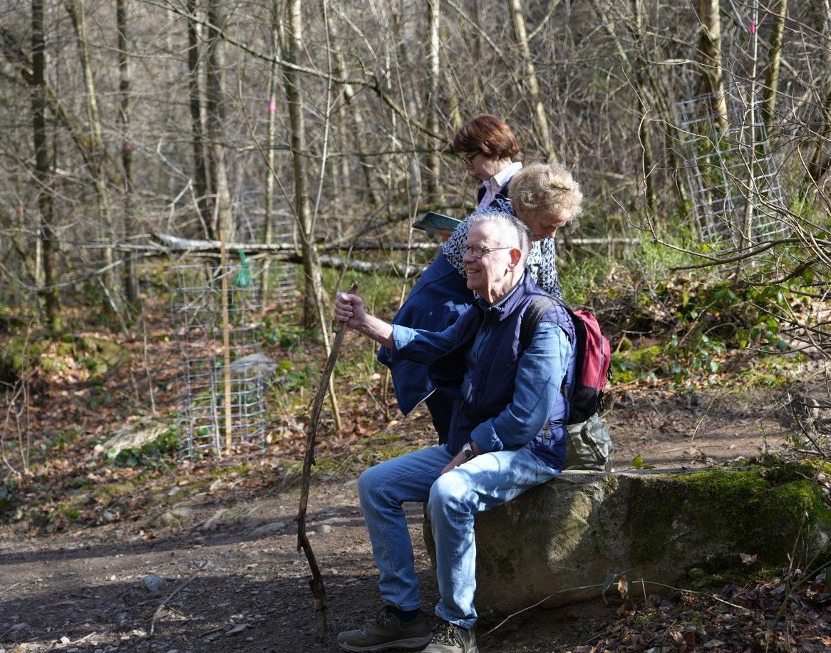 Drie mensen zitten tijdens een wandeling op een betonnen richel in een bosrijk gebied.