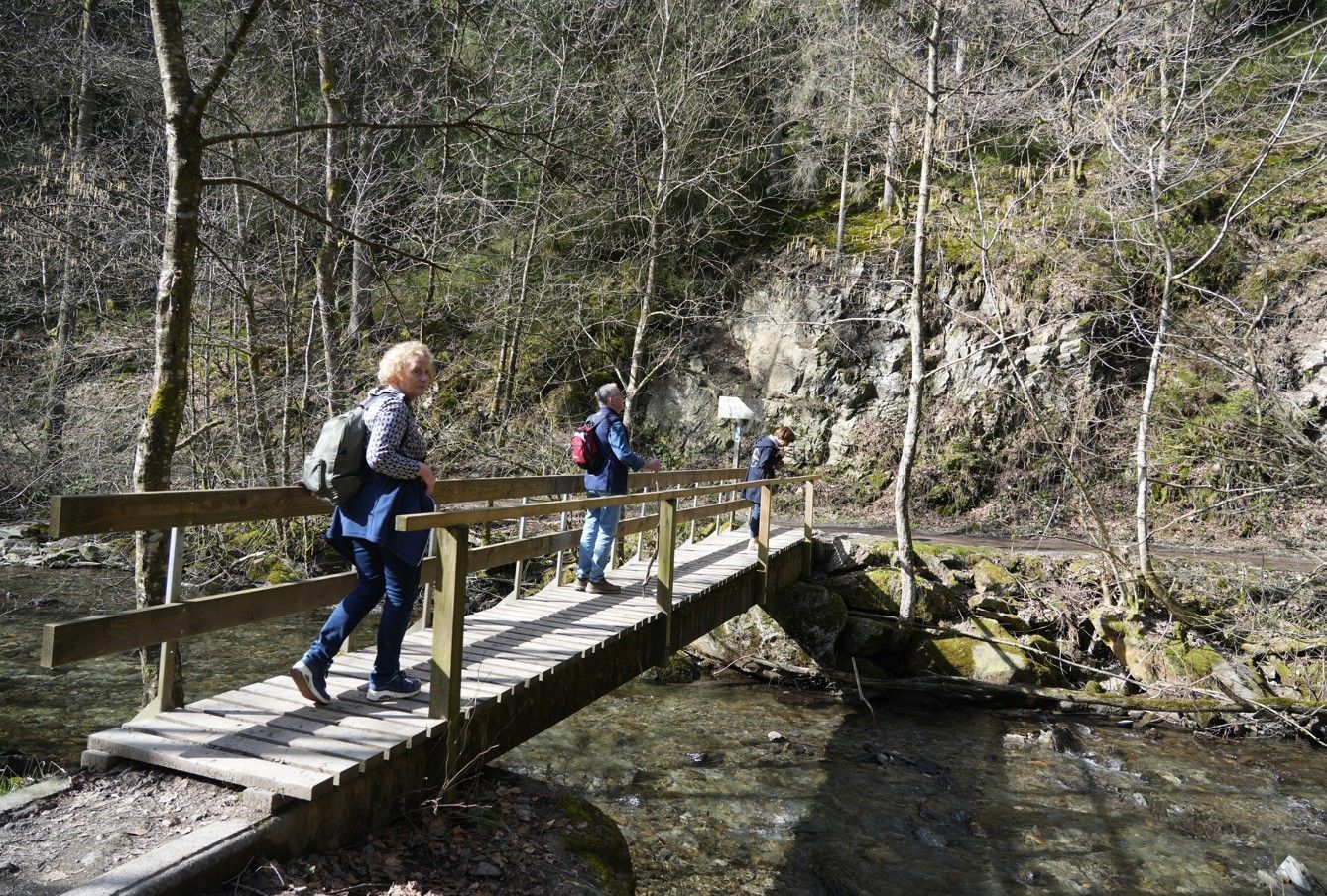 Op een zonnige dag lopen drie wandelaars over een houten bruggetje over een beekje in een bosrijk, rotsachtig landschap.