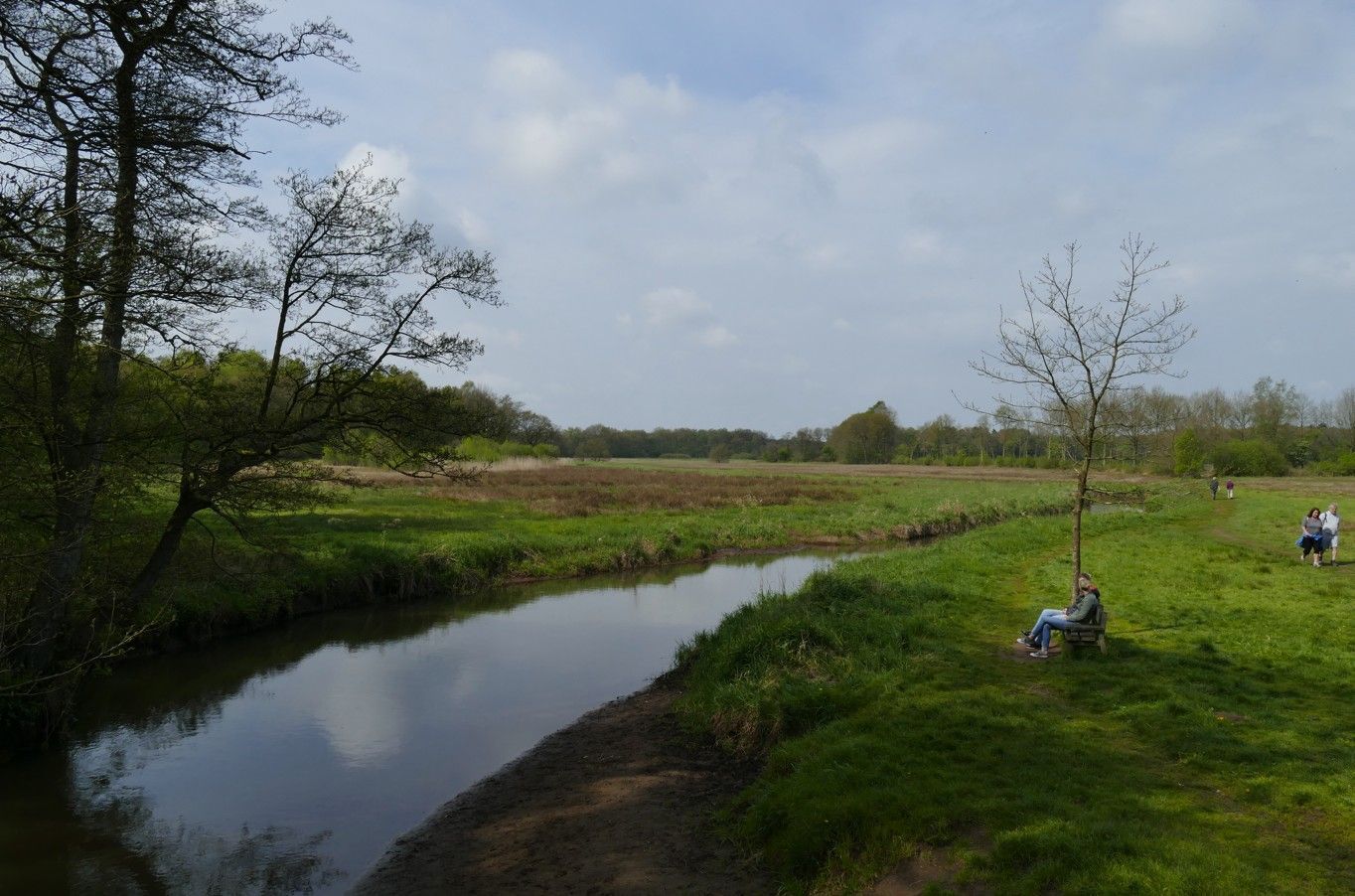 Een klein beekje stroomt door een grasveld; twee mensen ontspannen op de oever onder een kleine boom.