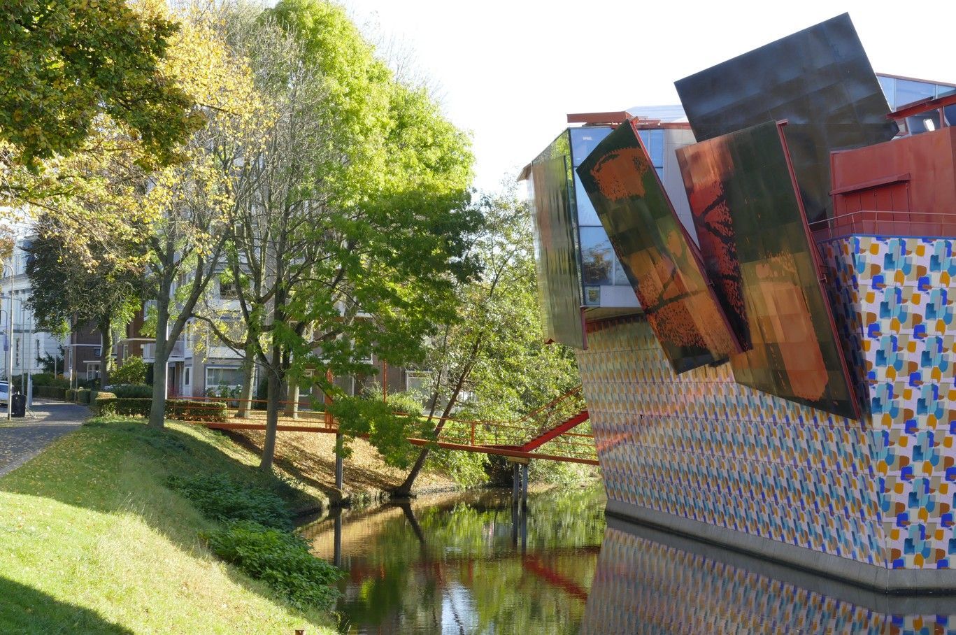 Een kanaal met bomen, een grasrijke oever en een modern gebouw met hoekige, kleurrijke tegels en metalen sculpturen.