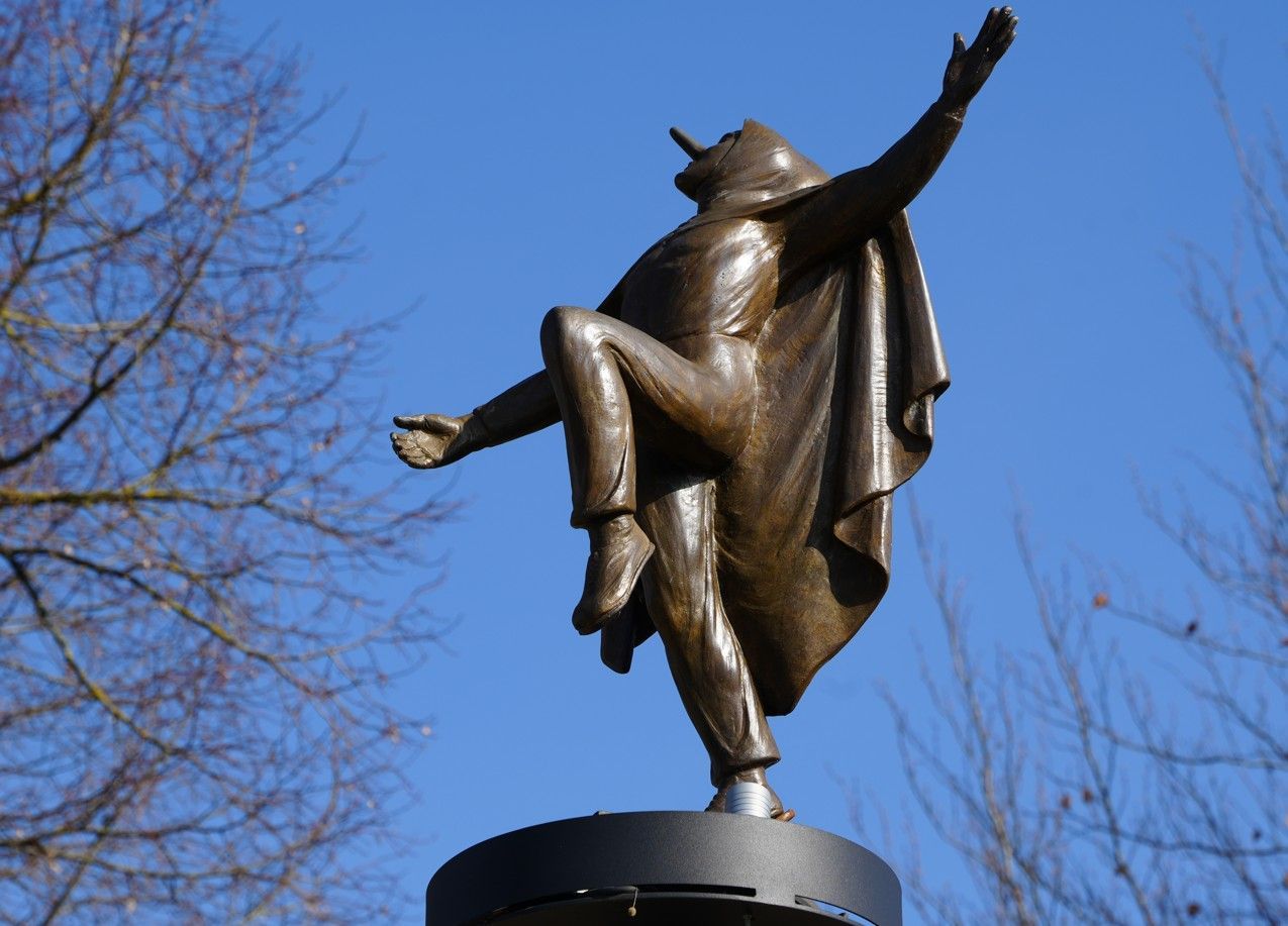Een bronzen beeld van een persoon in een cape, afgebeeld midden in een sprong met uitgestrekte armen, tegen een helderblauwe hemel.