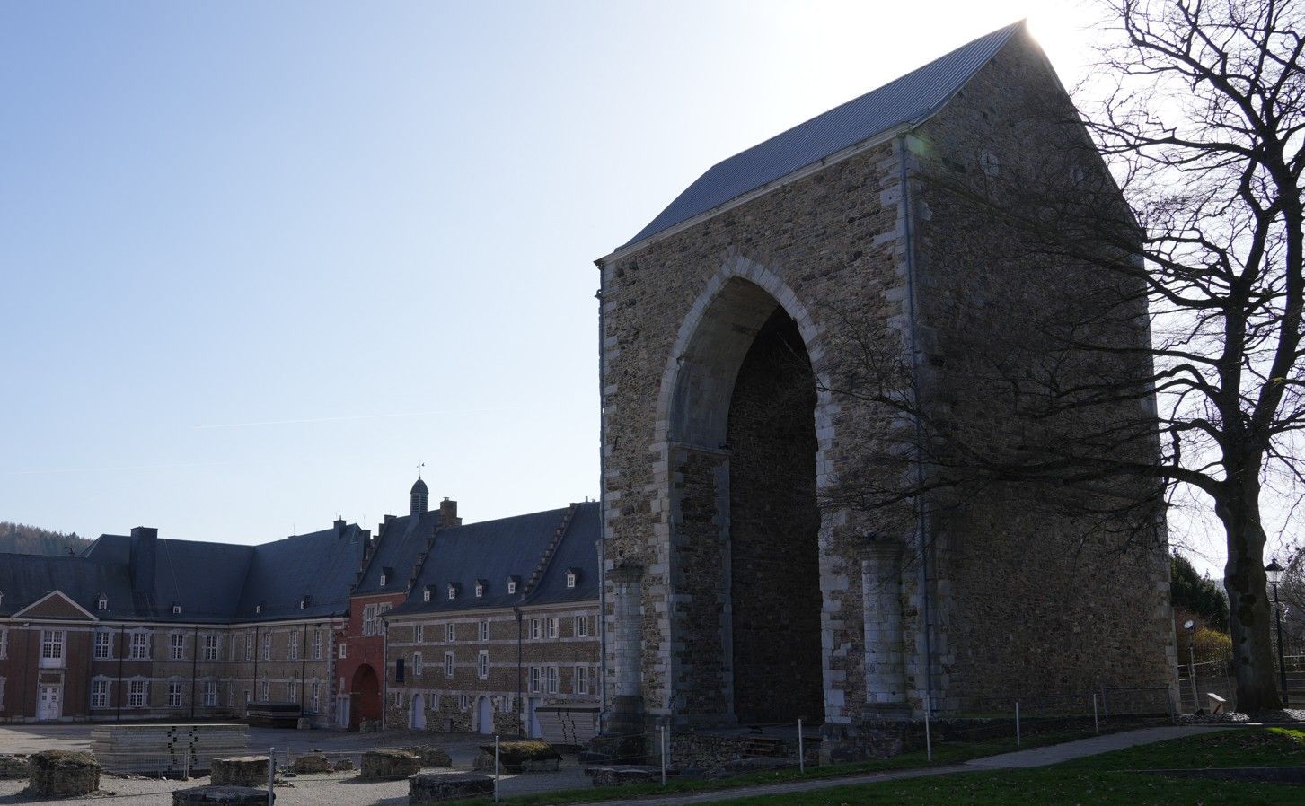Een stenen ruïne met bogen staat naast een groot, meerlaags bakstenen gebouw onder een helderblauwe hemel.