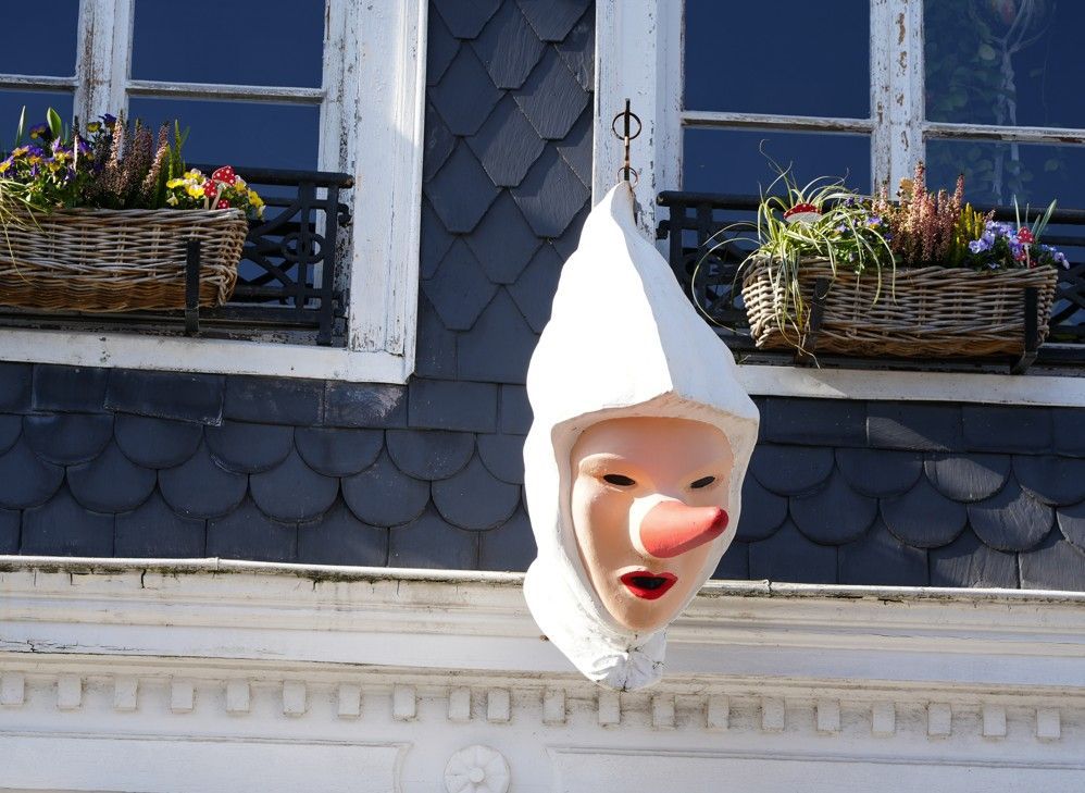 Een masker met een spitse neus en een kap hangt aan de muur tussen ramen die versierd zijn met bloembakken.