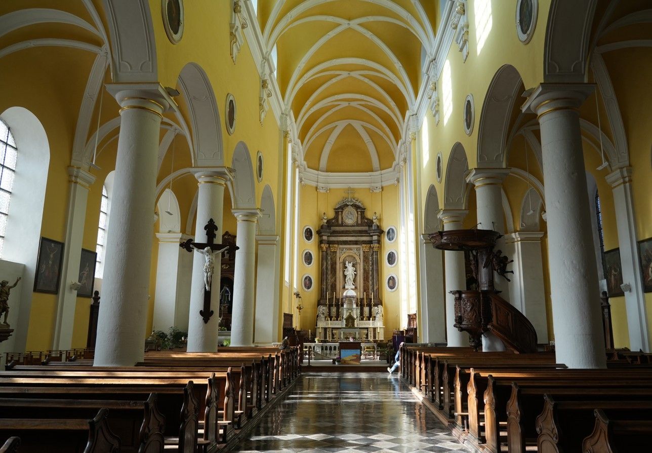 Interieur van een kerk met gele muren, witte zuilen, rijen houten kerkbanken en een altaar aan het uiteinde.