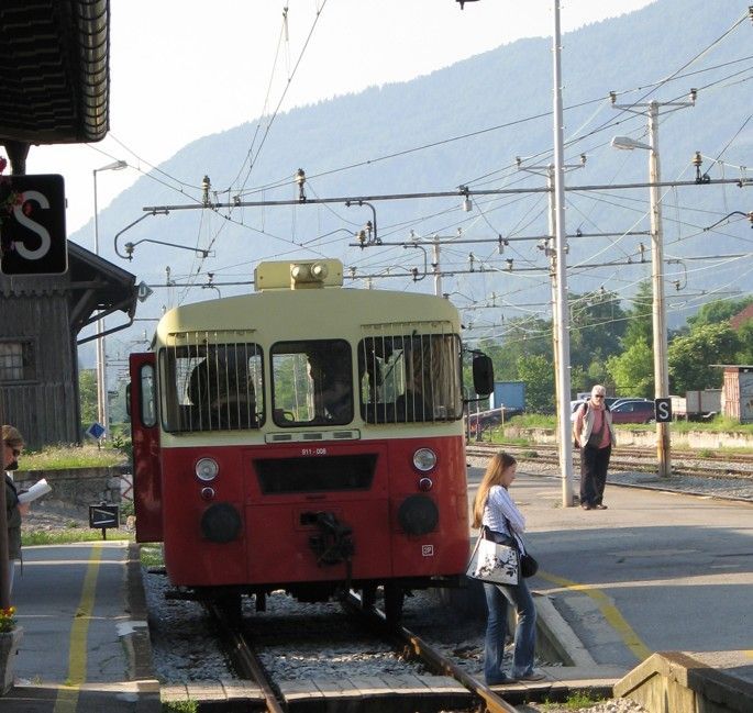 Een rood-crèmekleurige trein staat op een station. Een vrouw stapt uit. Een man staat er vlakbij. Op de achtergrond zijn bergen te zien.