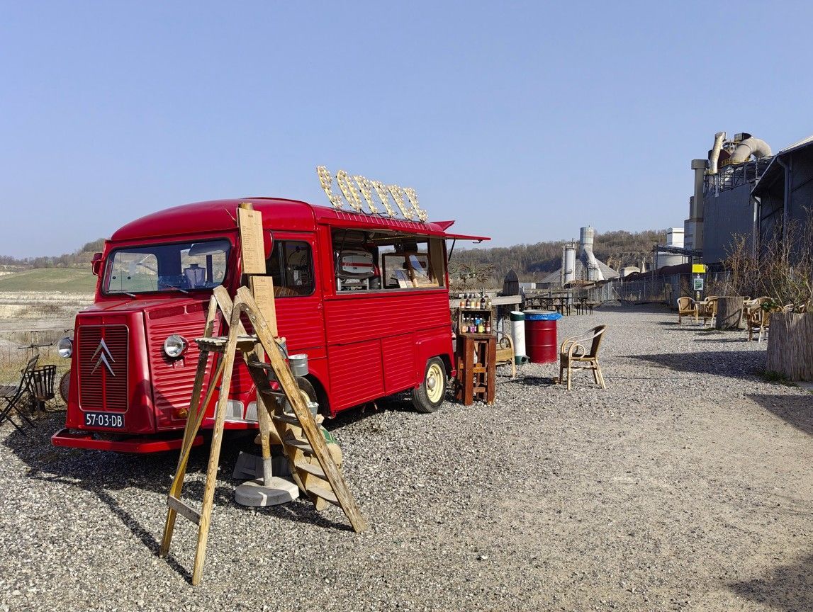 Een felrode foodtruck staat geparkeerd op een grindterrein in een landelijke, industrieel ogende omgeving.