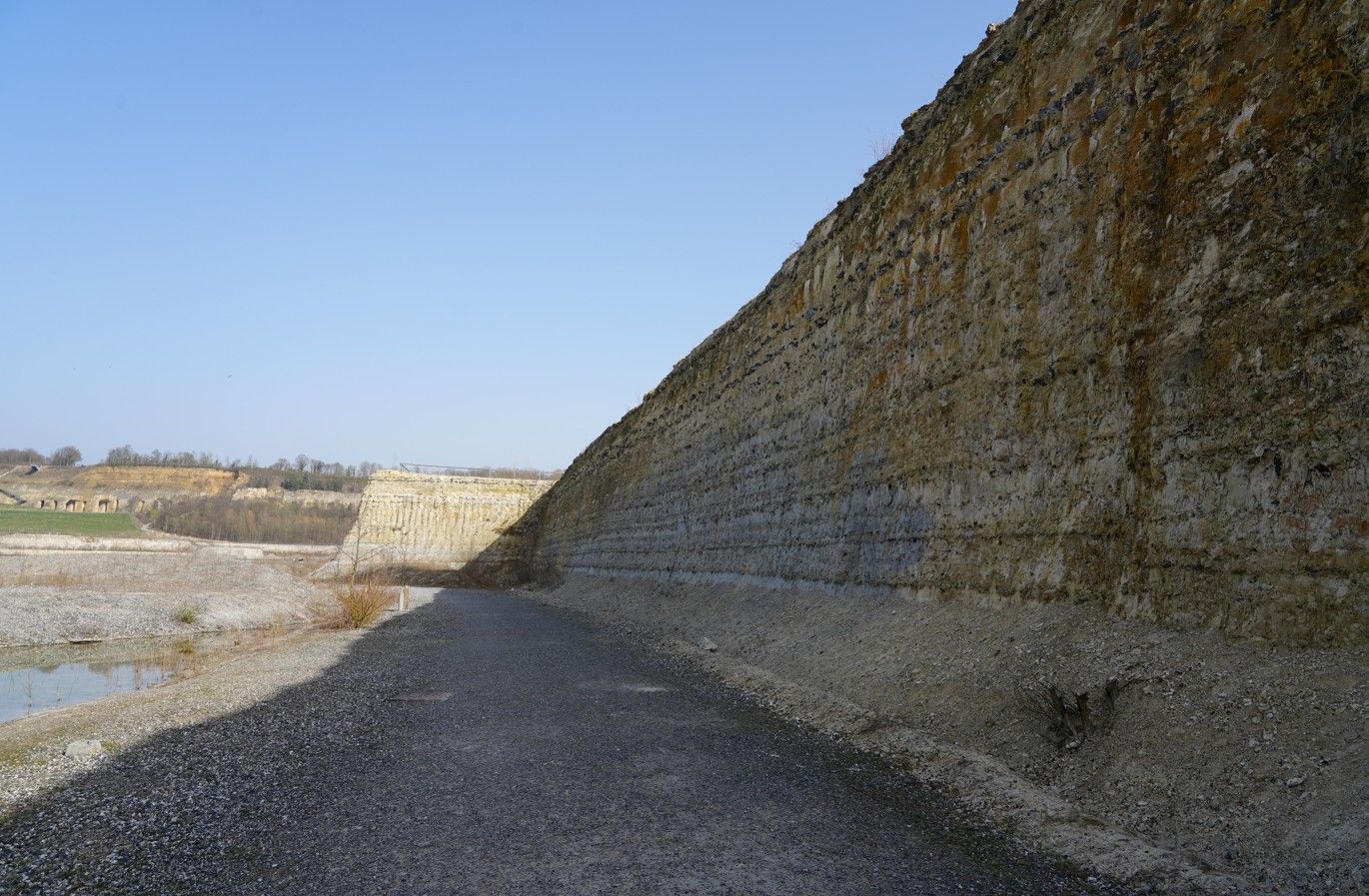 Een grindpad loopt langs een hoge, verweerde stenen wal onder een helderblauwe hemel.