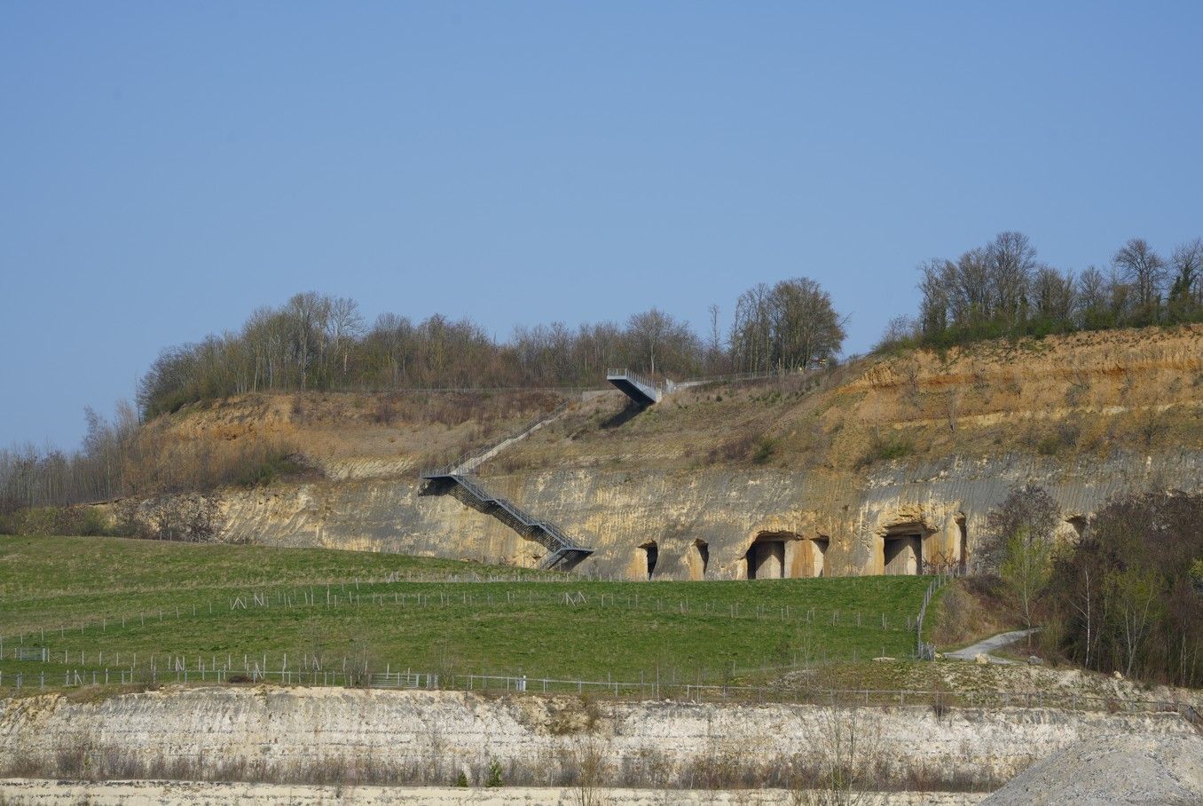 Een drone vliegt langs een met gras begroeide heuvel met een rotswand met verschillende grotachtige openingen.
