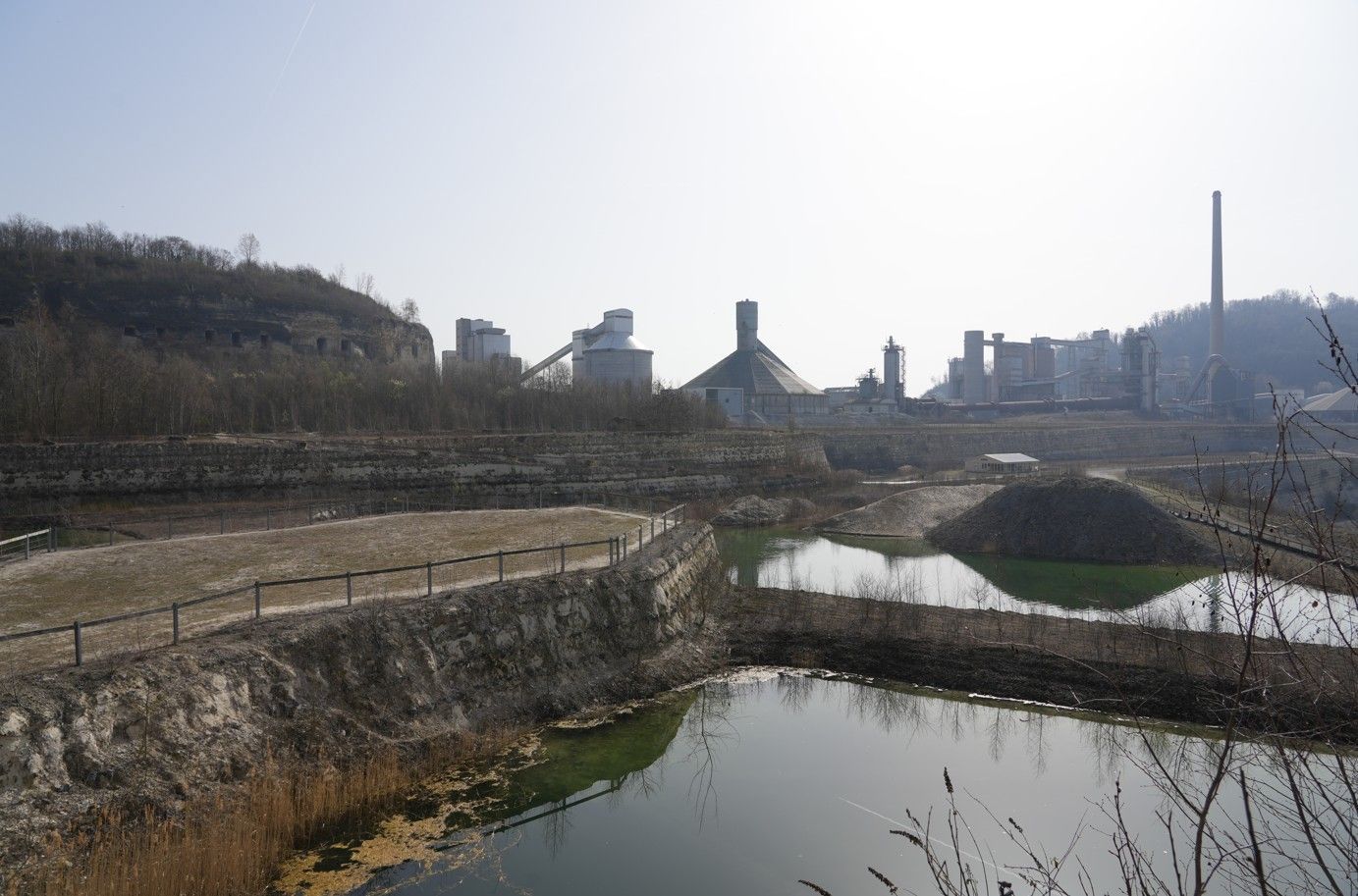 Een cementfabriekcomplex torent boven een trapsgewijs, rotsachtig steengroevelandschap uit, met spiegelend water.