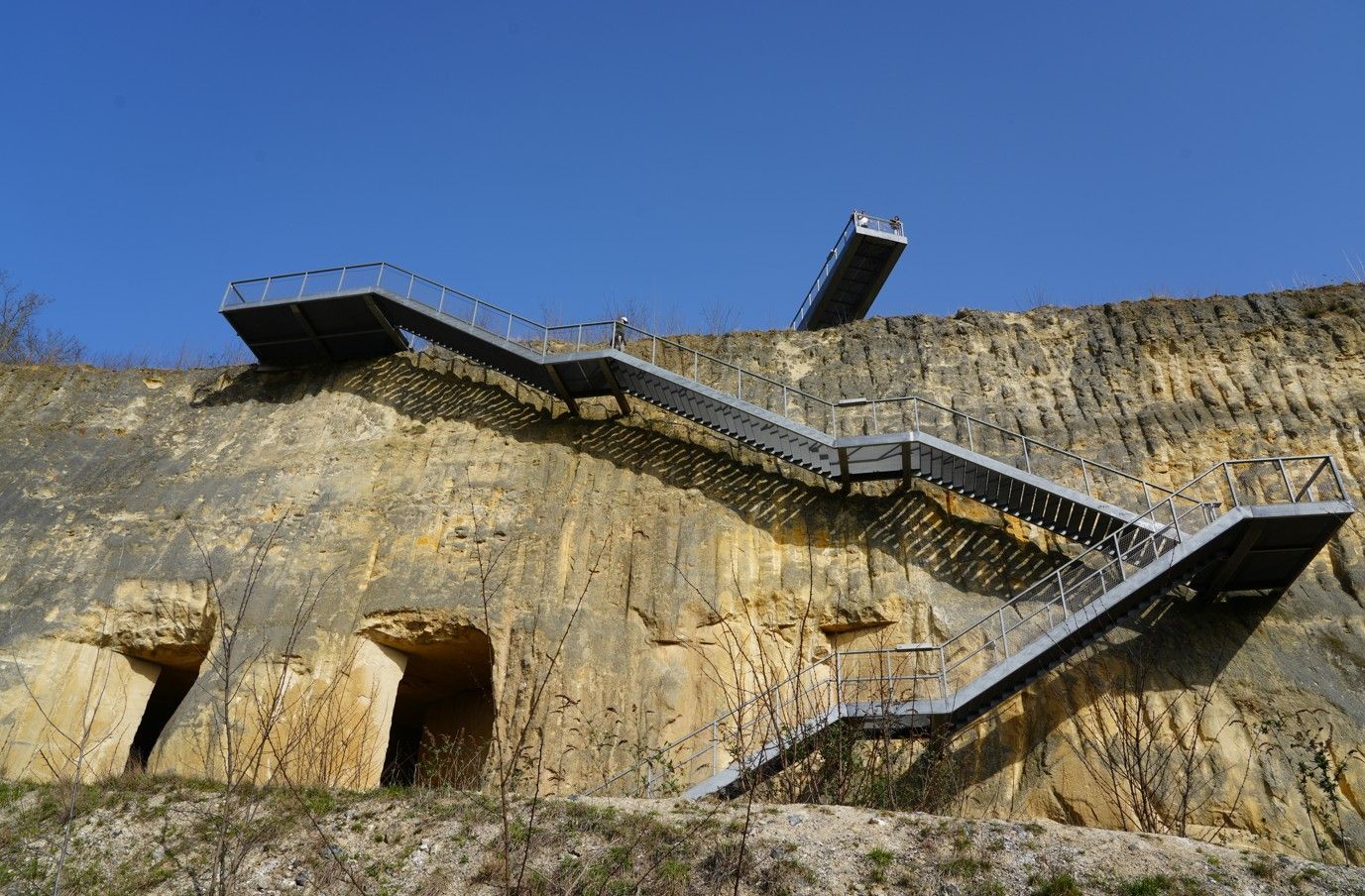 Een metalen trap slingert zich omhoog langs een ruige, lichtgekleurde kalkstenen klif naar een observatieplatform.