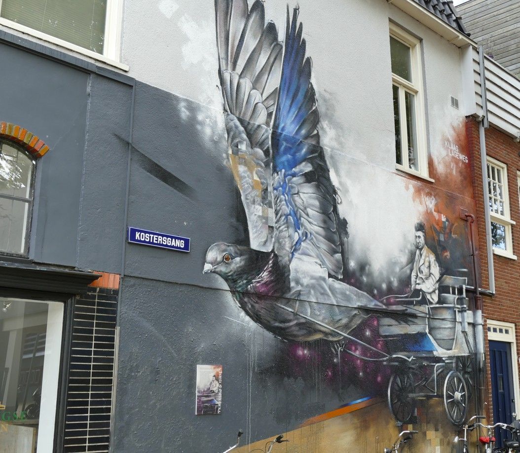 Muurschildering van een grote vogel met gespreide vleugels die een koets trekt. Kleuren: grijs, blauw en wit. Aangebracht op de buitenkant van een gebouw.