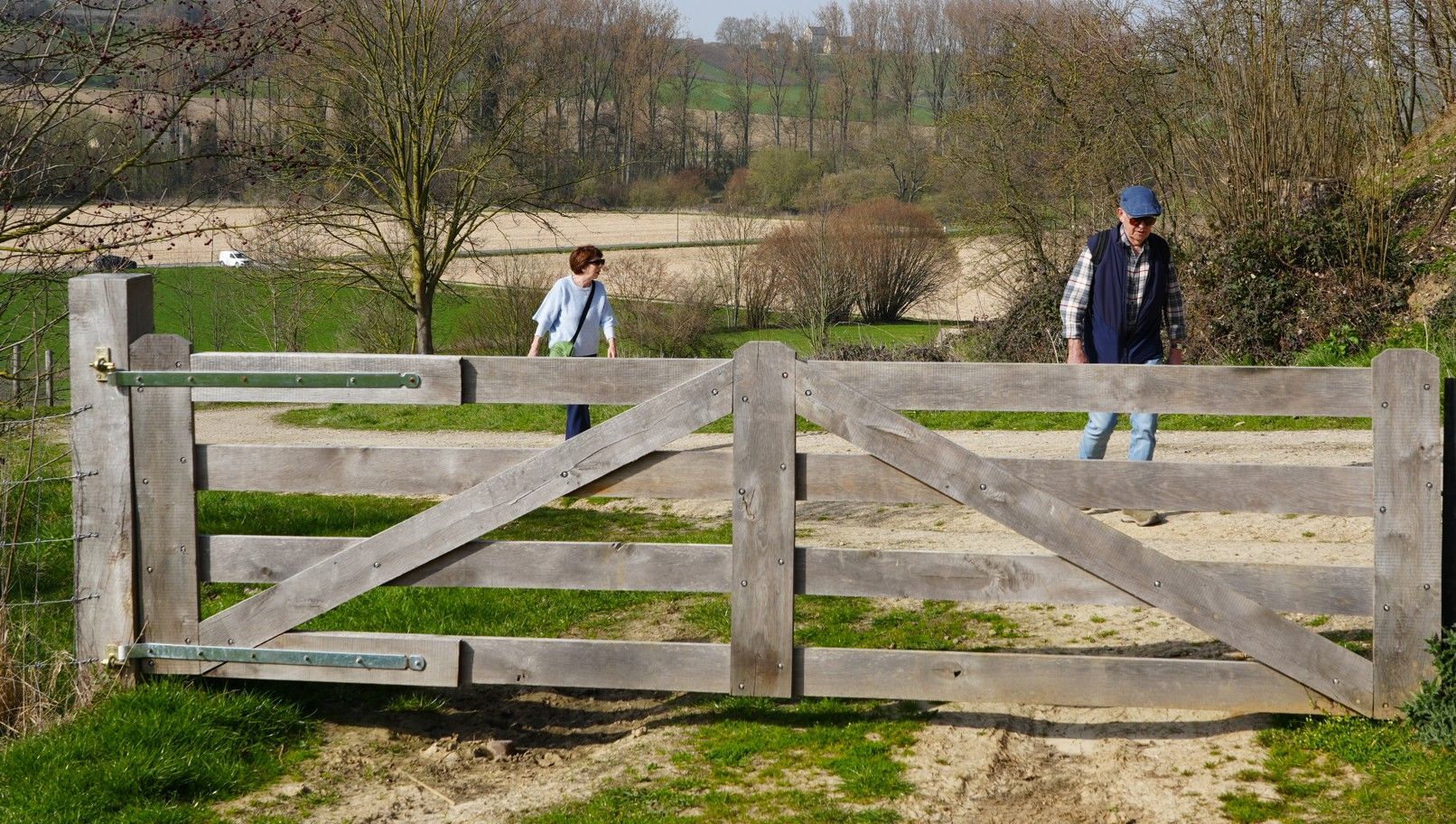 Op de voorgrond staat een houten boerenpoort, terwijl twee mensen over een zandpad lopen in een landelijke buitenomgeving.