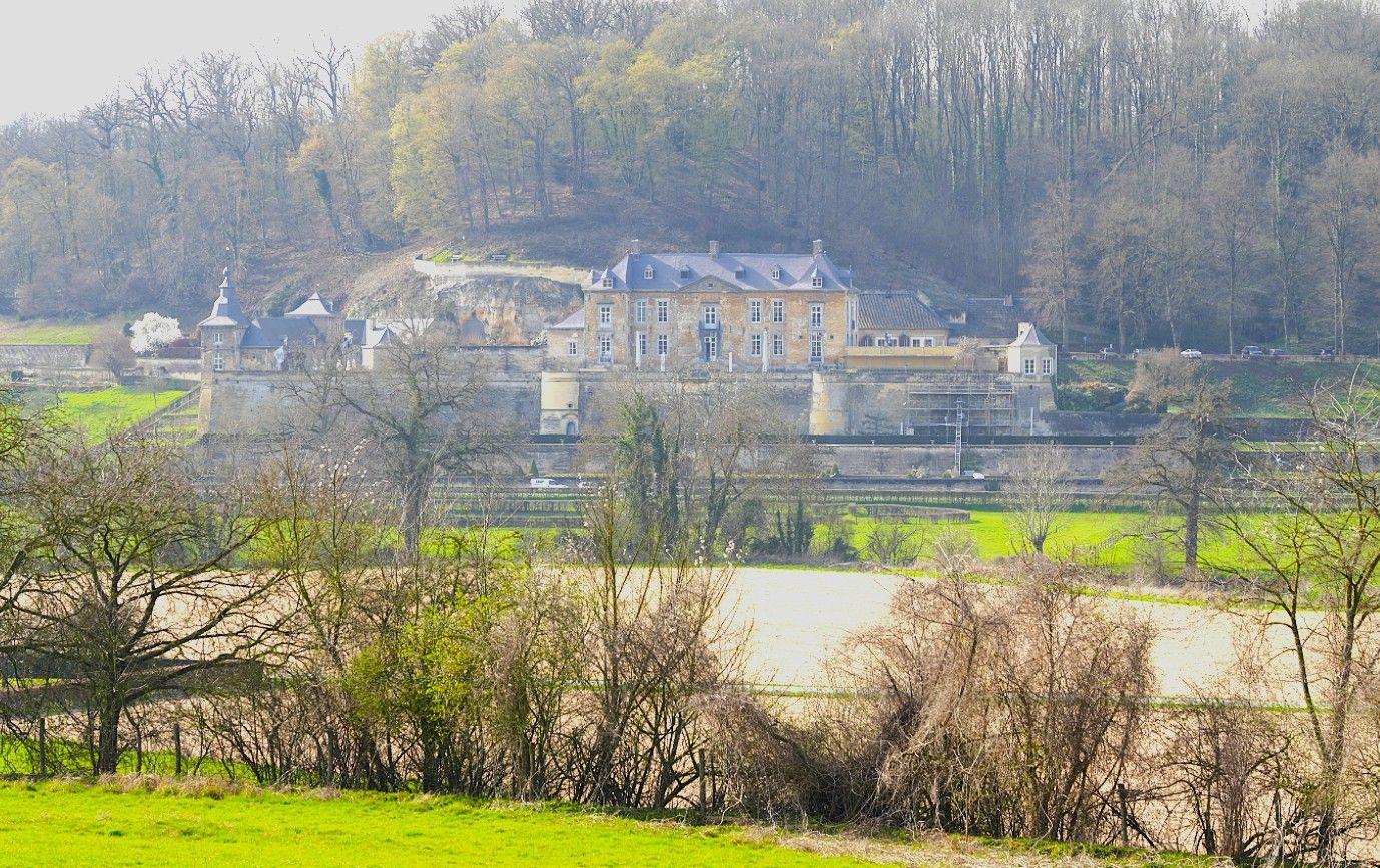 Op een heuvel staat een stenen kasteel met een grijs dak, dat uitkijkt over een groene vallei en omringende bomen.