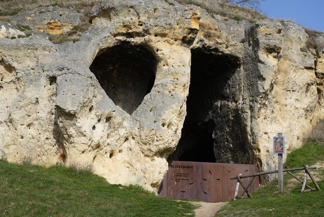 Een geërodeerde kalkstenen rotswand met een hartvormige holte en een donkere, rechthoekige grotingang, met een metalen hek.