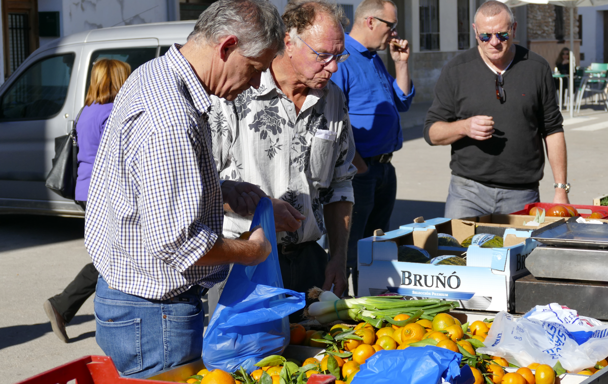 Mensen op een openluchtmarkt bekijken groenten en fruit. Sinaasappels en andere groenten liggen uitgestald.