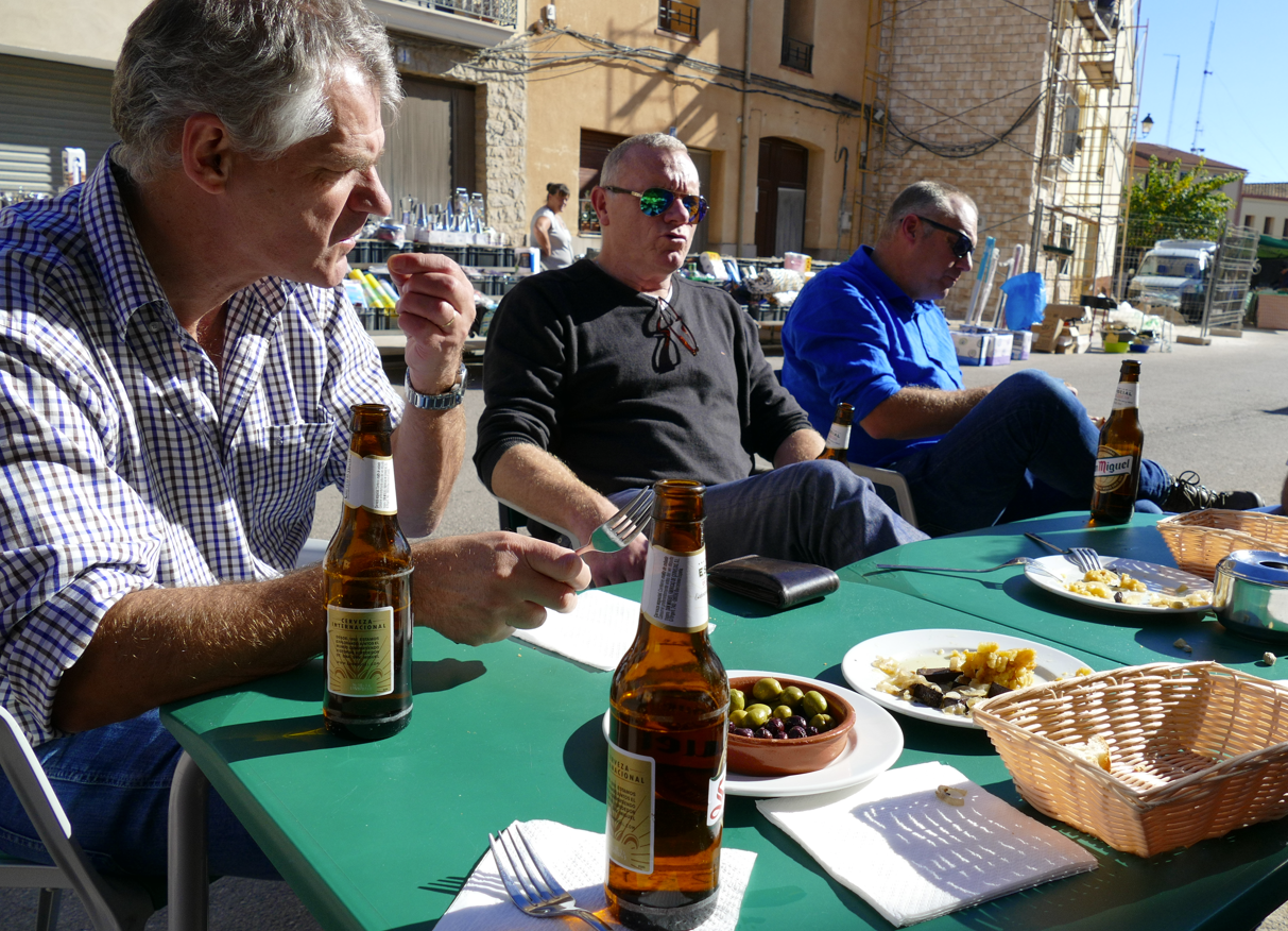 Drie mannen aan een tafeltje buiten, met bier, olijven en eten. Een zonnige dag op een Europees stadsplein.