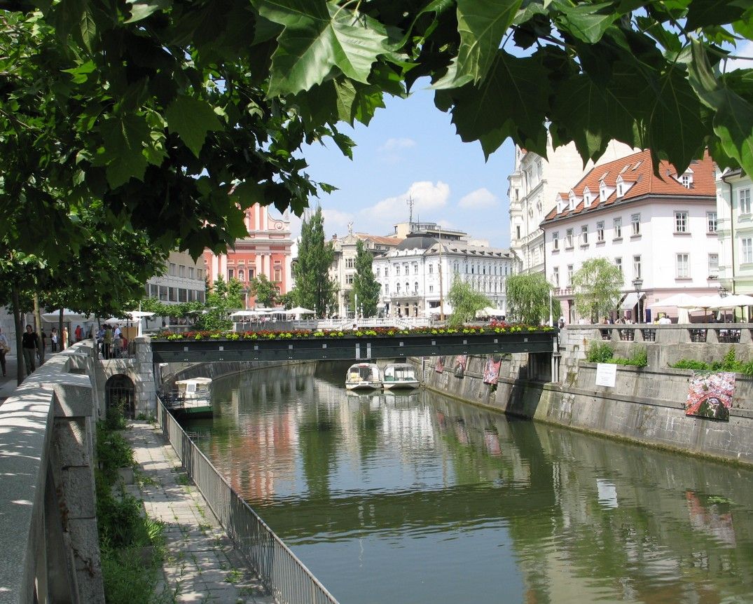 Kanaal in Ljubljana, Slovenië, met brug, boten en gebouwen op de achtergrond onder een zonnige hemel.