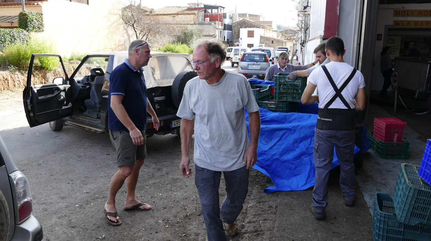 Mannen lossen kratten uit een voertuig in een straatbeeld. Een andere man loopt in de richting van de camera.