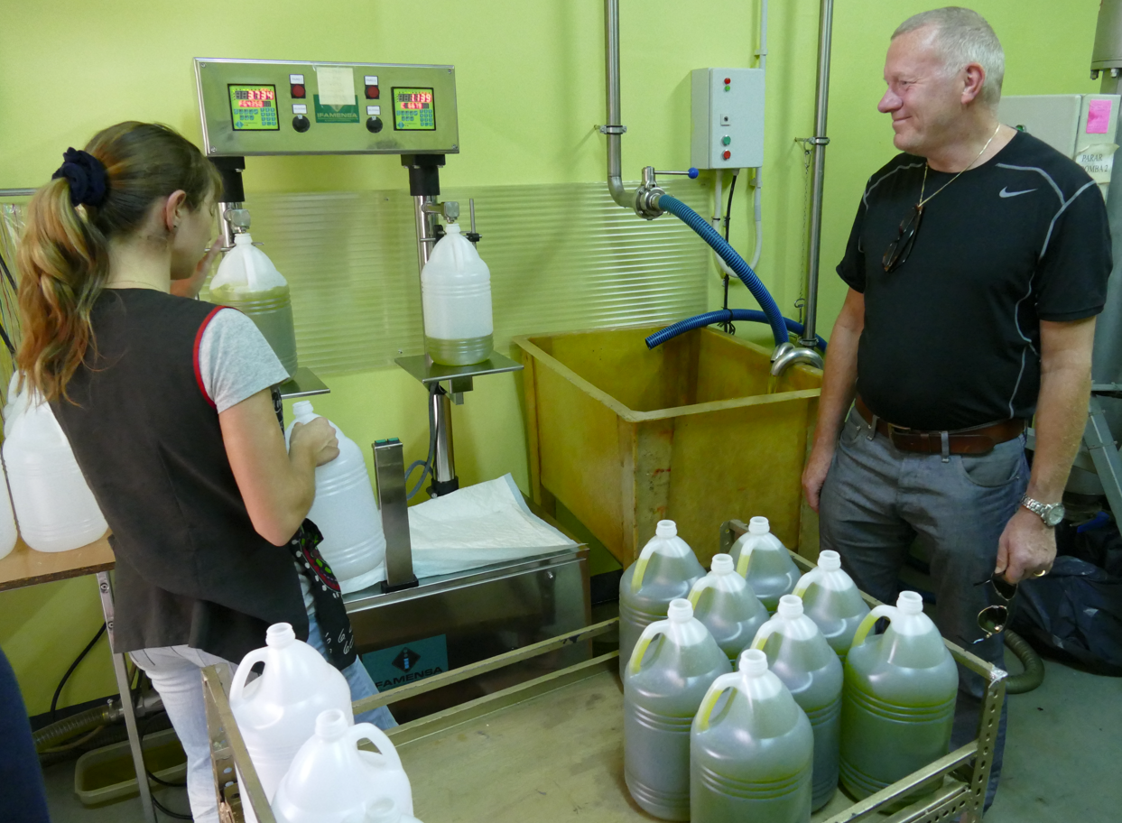 Twee mensen zijn bezig met het bottelen van vloeistof in een fabriek. De ene vult containers, de andere kijkt toe. Er zijn groene en witte containers aanwezig.