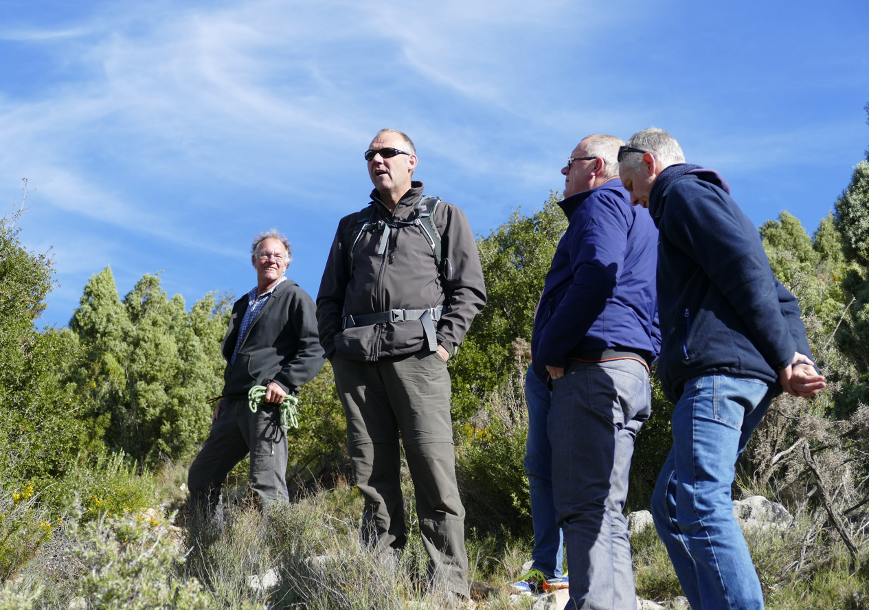 Vier mannen in vrijetijdskleding staan ​​buiten en kijken naar iets onder een blauwe hemel, met struiken en lage begroeiing op de voorgrond.