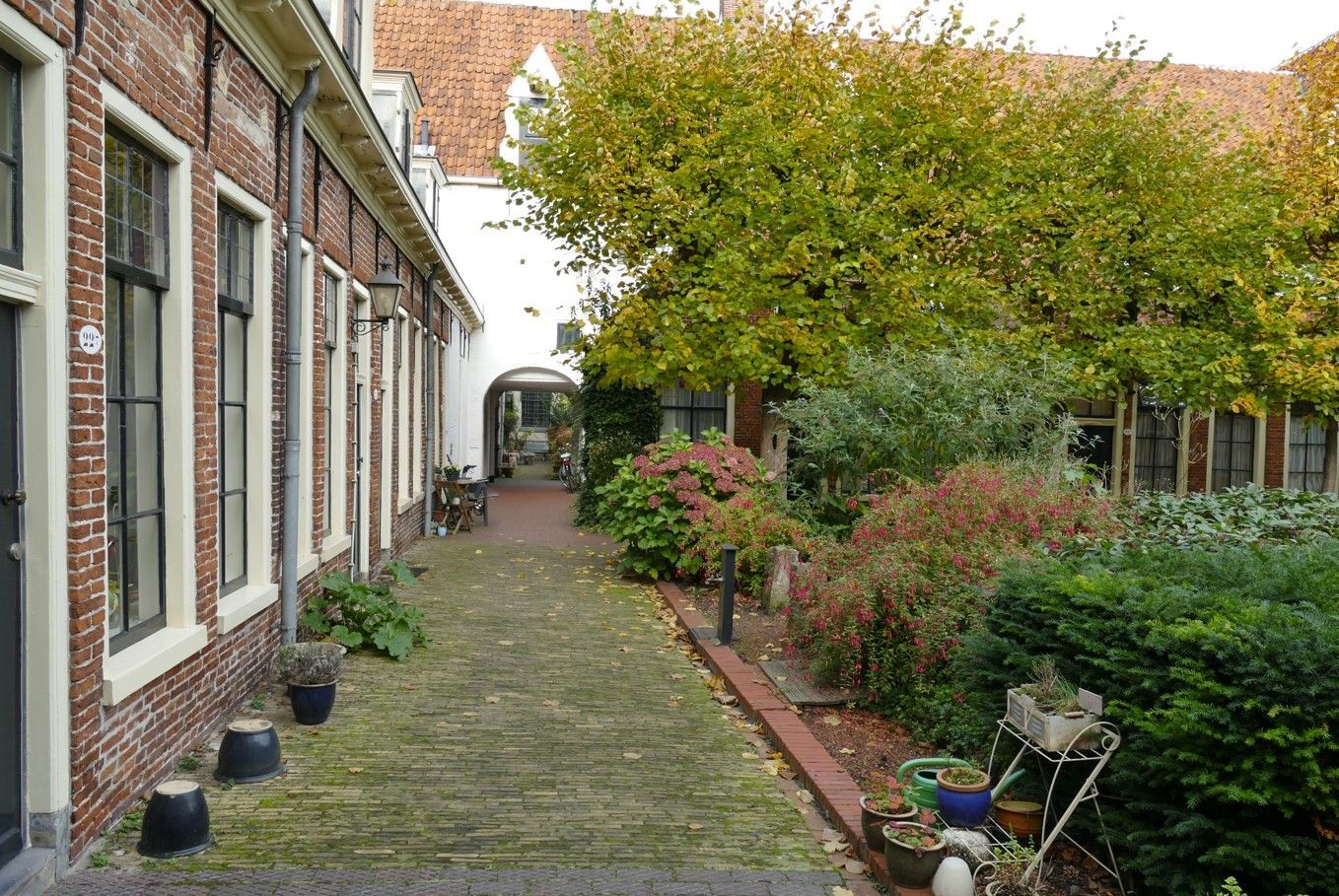 Bakstenen gebouwen met hoge ramen omringen een bakstenen pad dat naar een boog en groen leidt.