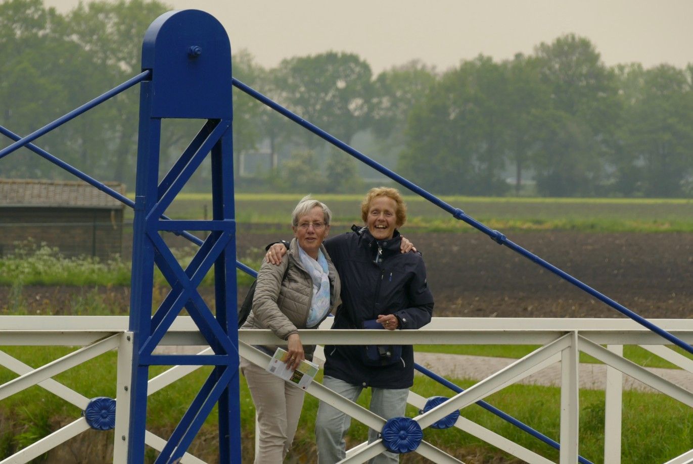 Twee mensen op een blauw-witte brug, lachend naar de camera, met een veld en bomen op de achtergrond.