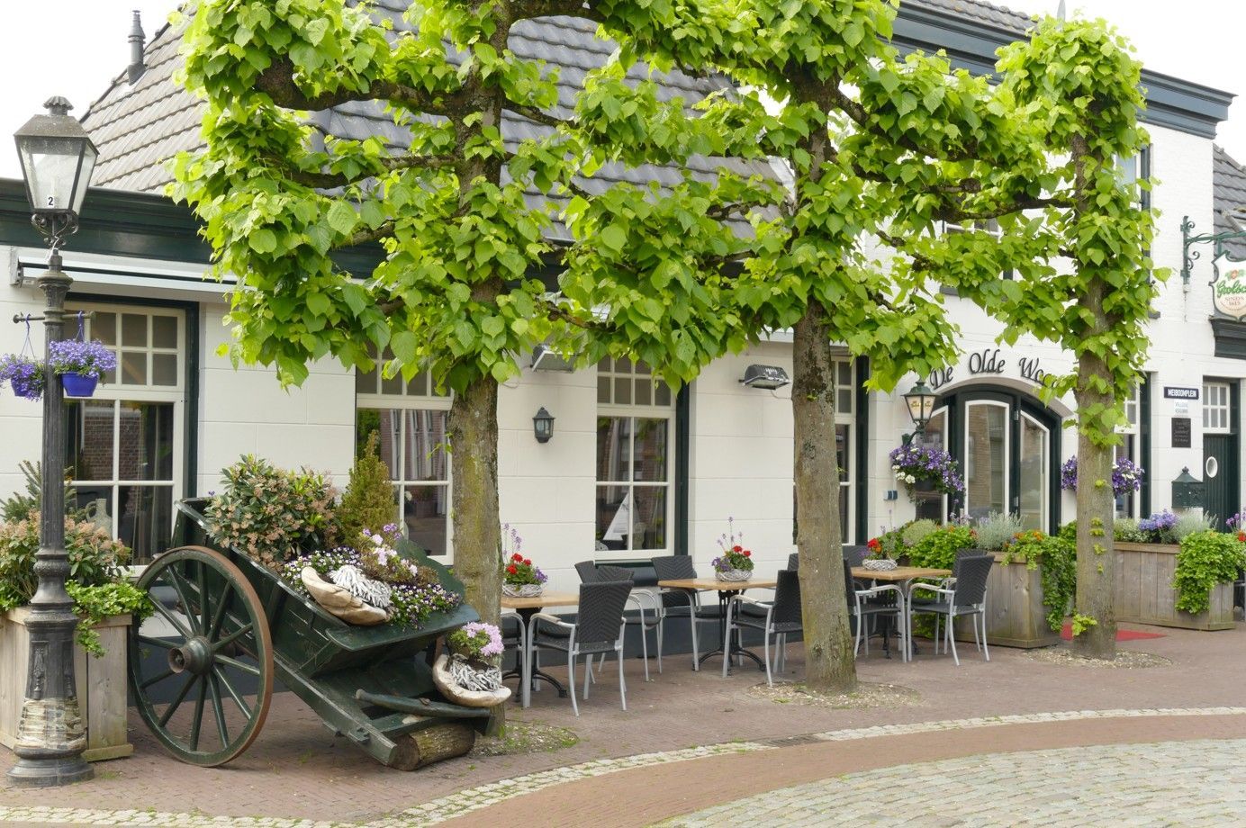 Wit gebouw met zitgelegenheid buiten onder gesnoeide bomen, kar met bloemen.