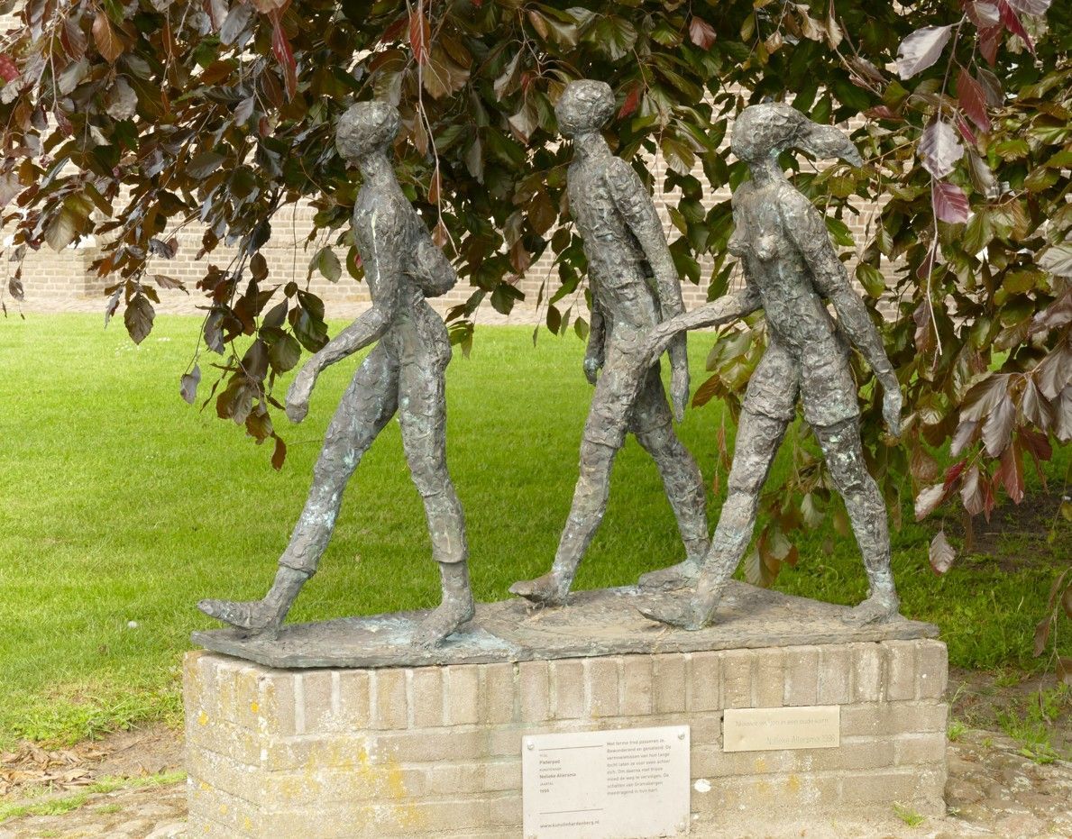 Bronzen sculptuur van drie figuren die op een stenen sokkel onder een boom lopen.