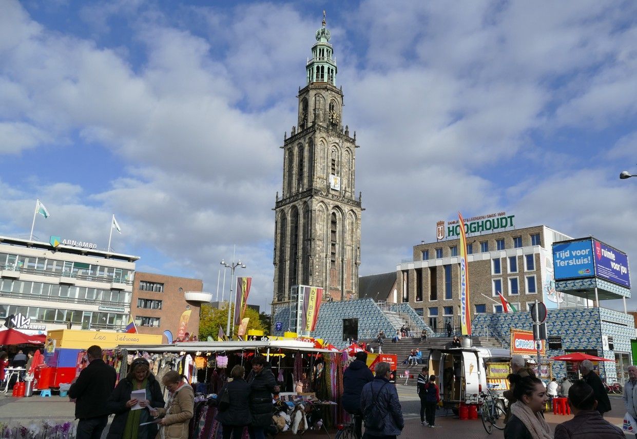 Marktplein met hoge toren, winkels en mensen onder een bewolkte hemel.