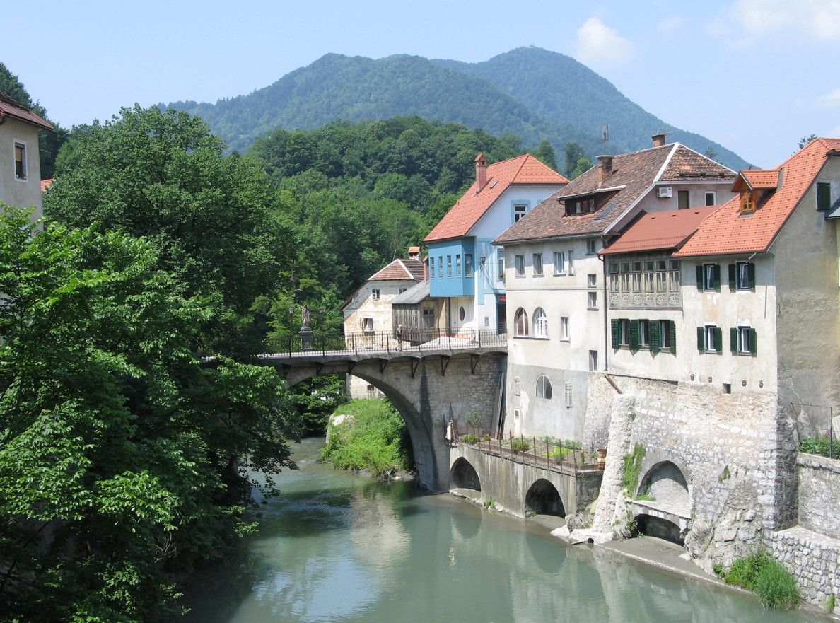 Stenen brug met huizen langs een rivier in een Europees dorp; groene bomen, blauwe lucht.