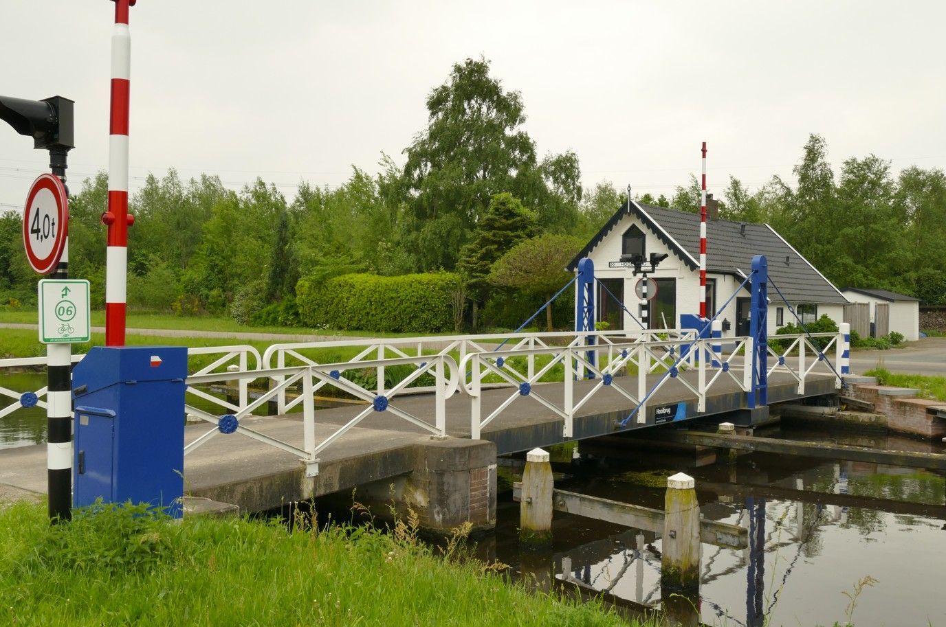 Een blauw-witte ophaalbrug over een kanaal met een klein bedieningshuisje, rood-wit gestreepte slagbomen en groen.