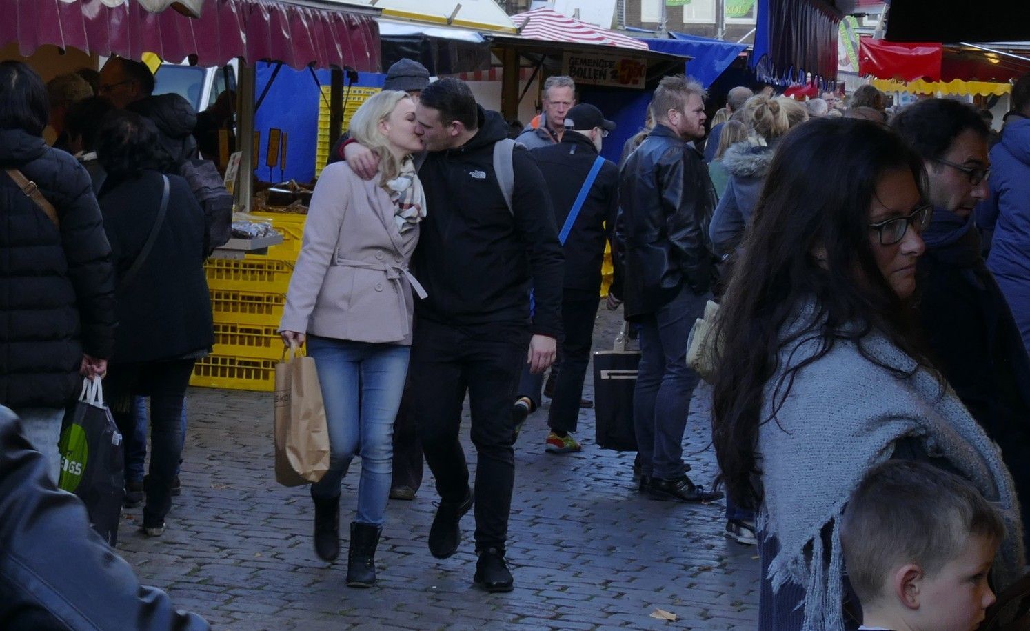 Een stel dat elkaar kust op een drukke openluchtmarkt; de vrouw draagt ​​een jas en jeans, de man een zwart jack.