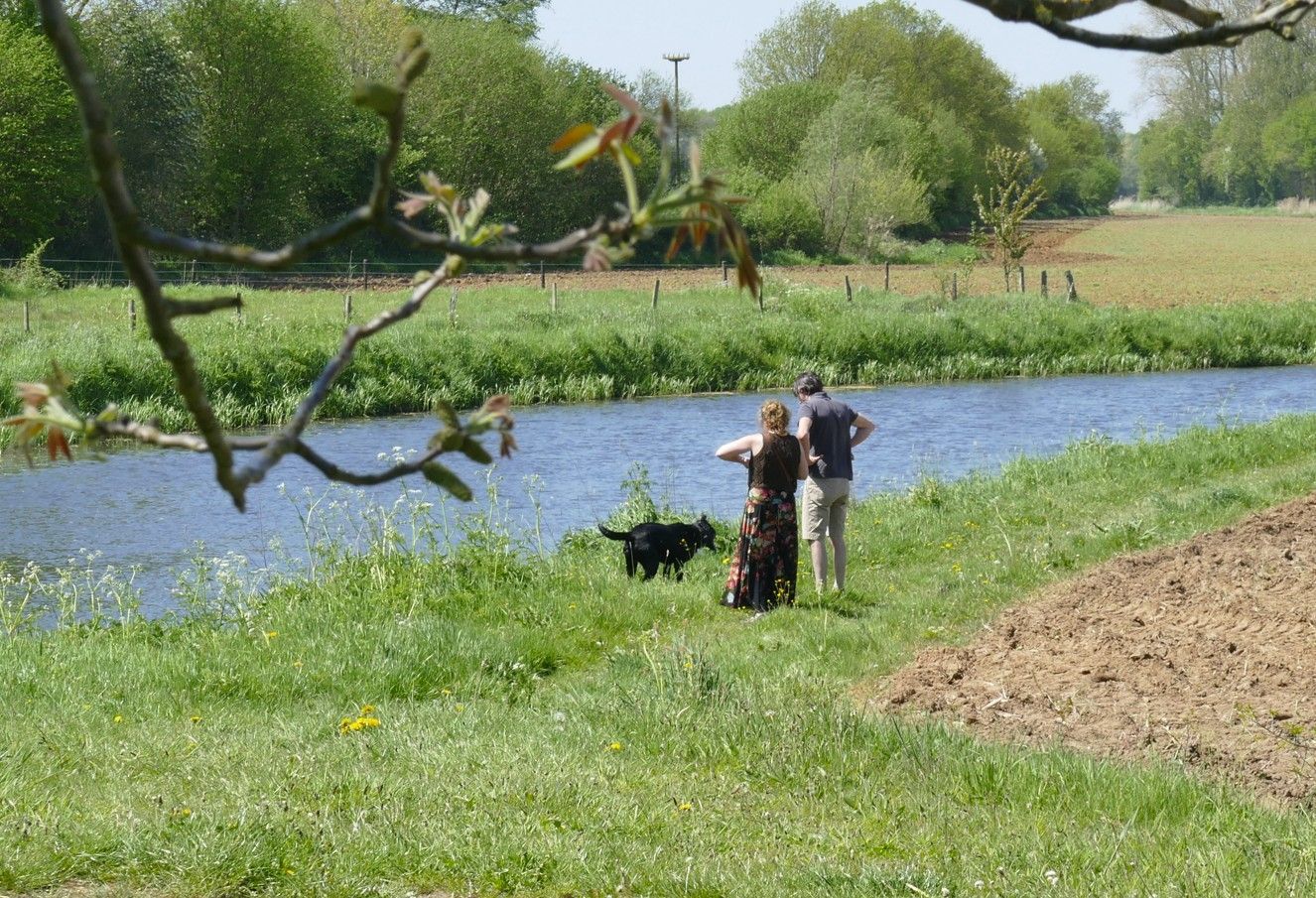 Mensen met een hond bij een rivier, staand op de grashelling. Bomen en velden op de achtergrond.