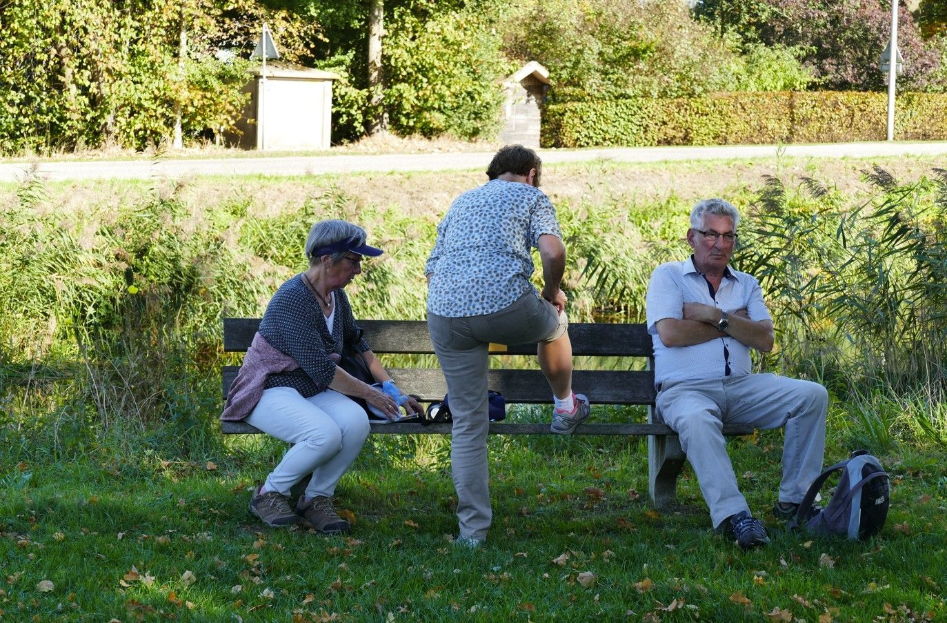 Drie mensen zitten en staan ​​op een houten bank in een grasveld in een park op een zonnige dag.
