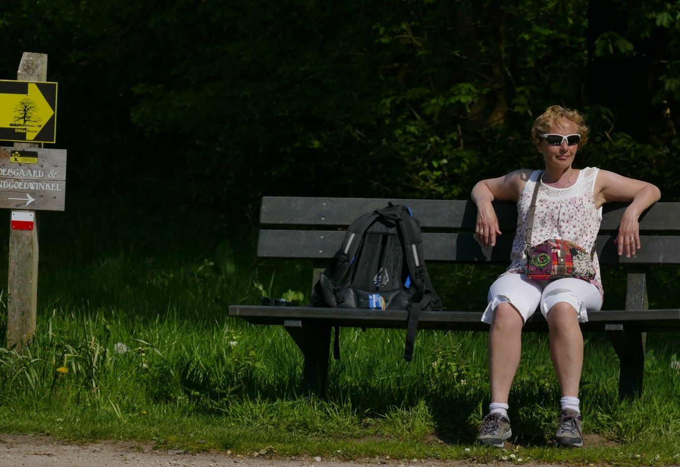 We rusten uit op een bankje bij een wandelpadmarkering in een bosrijk gebied. Naast haar ligt een rugzak.