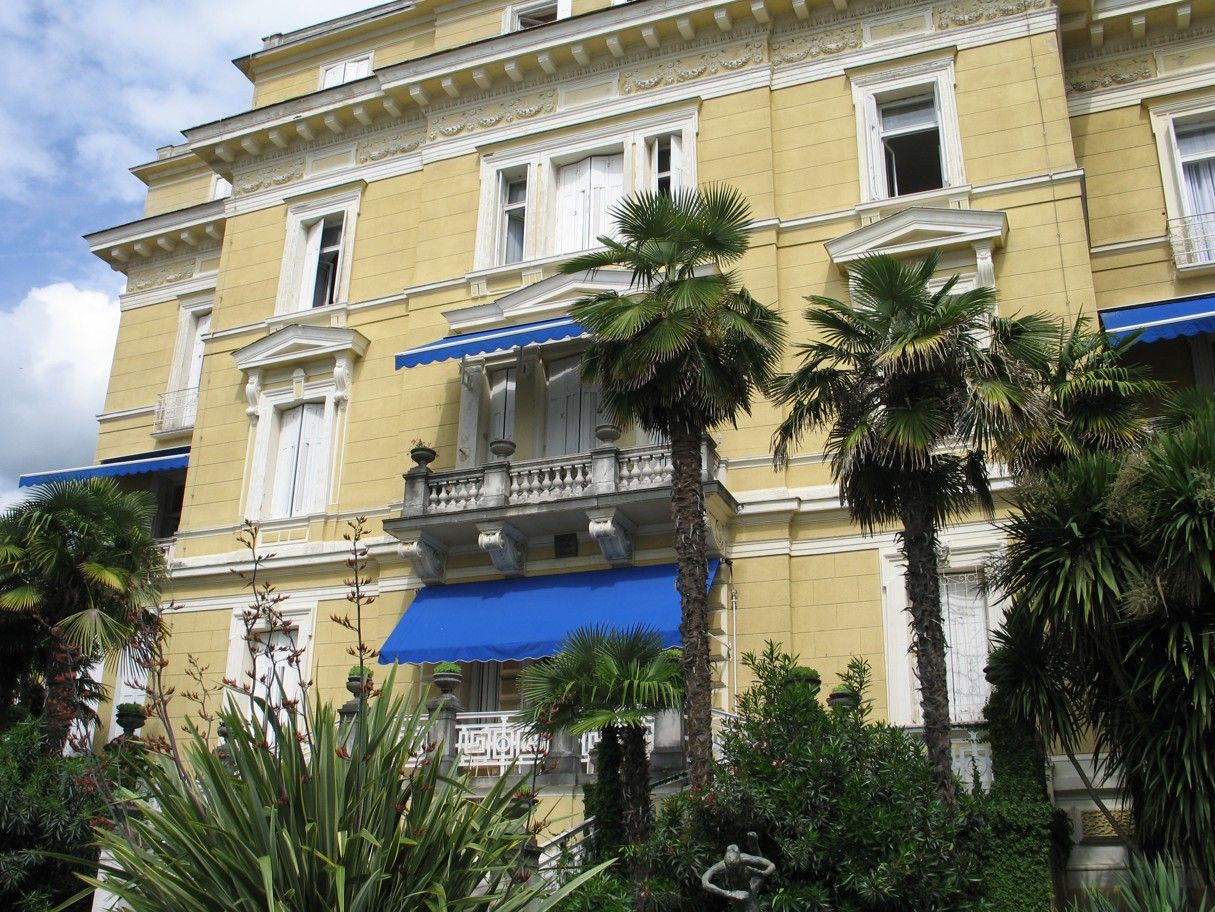 Geel gebouw met witte accenten, blauwe luifels en palmbomen.